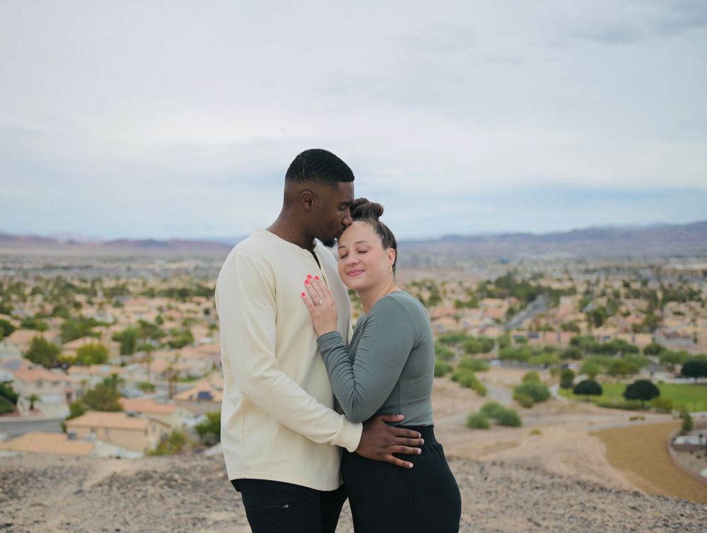 A couple embraces closely on a rocky desert hilltop overlooking a sprawling suburban neighborhood and distant mountains under a cloudy sky.