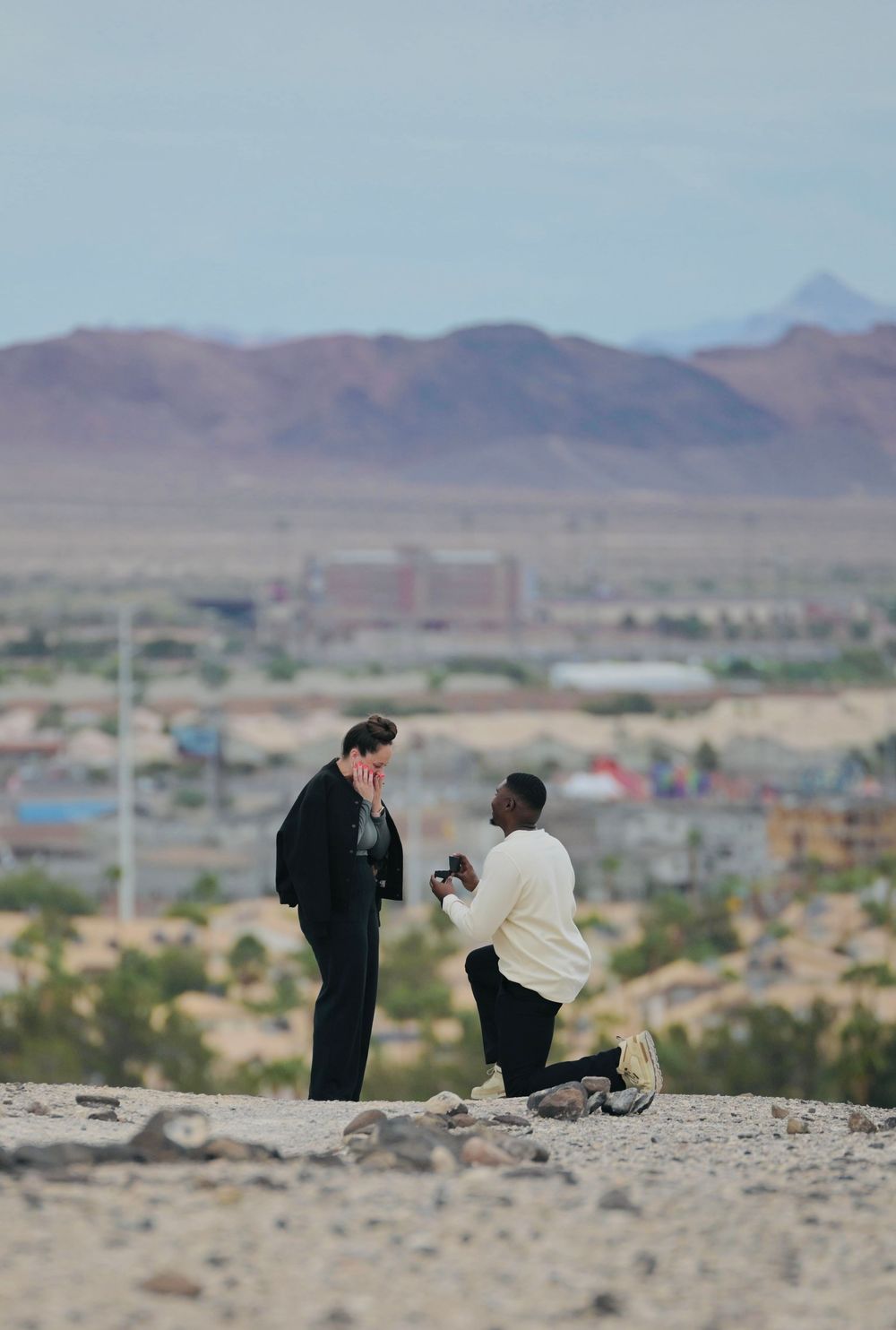 A man kneels on one knee proposing to his partner, who covers her face, on a rocky desert hilltop with mountains behind.