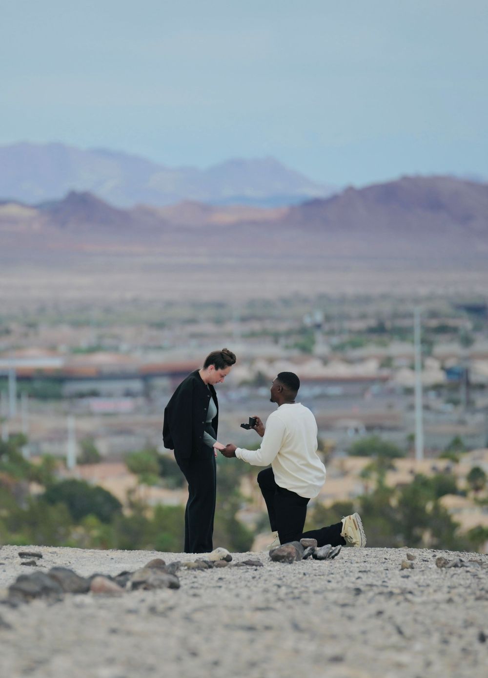 A man kneels on a rocky desert hill holding out a ring as he proposes to a woman, with purple mountains in the distance.