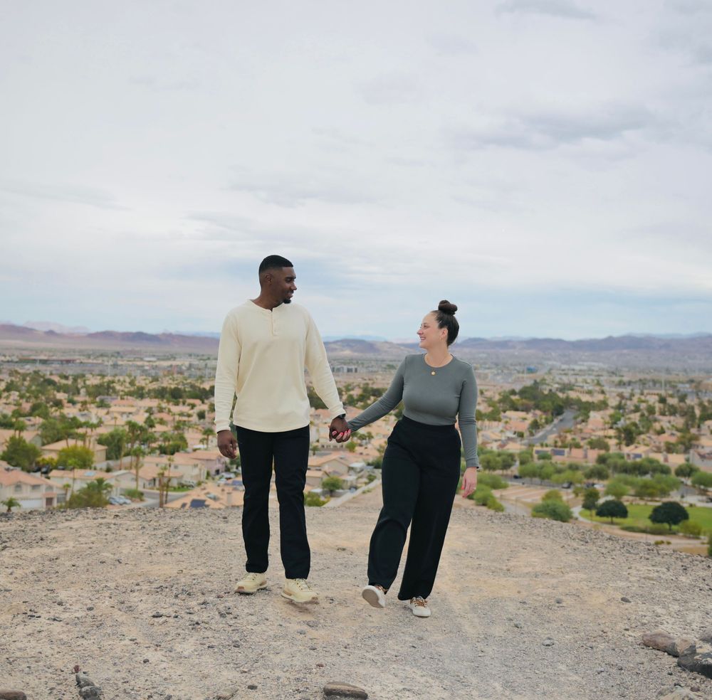 Smiling couple holding hands while walking along a rocky desert hilltop with a sweeping view of a suburban valley and mountains.