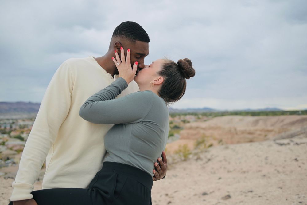 Couple in casual sweaters kissing and embracing outdoors on a rocky desert landscape under a cloudy sky.