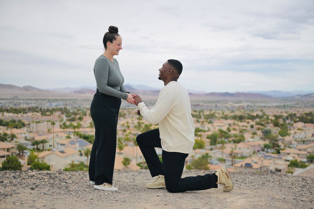 Man kneeling on one knee proposing to a smiling woman on a desert hilltop overlooking a sprawling suburban valley and distant mountains.