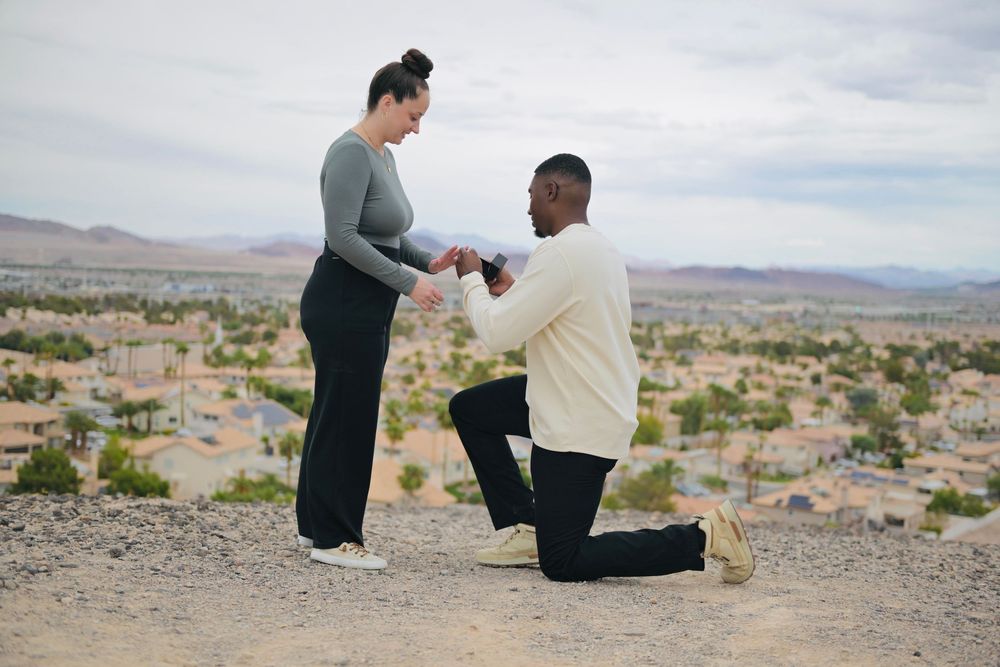 Man kneeling on one knee proposes to a smiling woman on a desert hilltop overlooking a sunlit suburban valley and distant mountains.