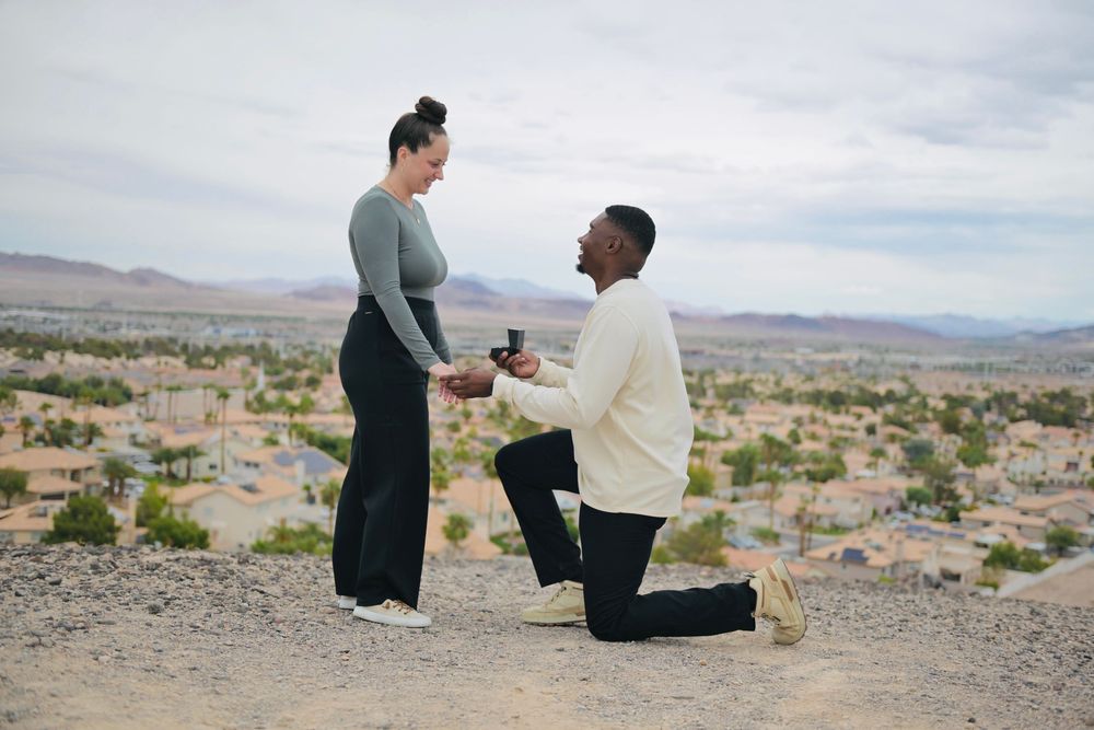 Man kneeling on one knee proposing to his smiling girlfriend on a desert hilltop overlooking a scenic valley at sunset.
