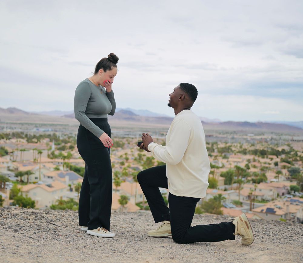Man kneels on one knee proposing to a woman covering her mouth in surprise on a desert hilltop overlooking a sunlit valley town.