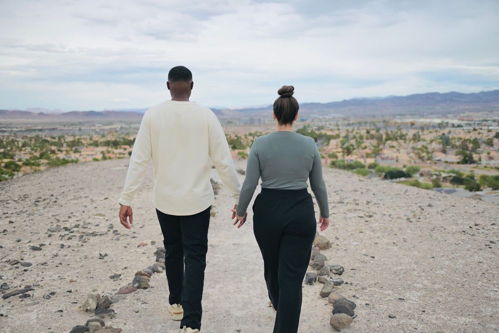 Couple holding hands walking away on a rocky desert trail toward distant mountains under a cloudy sky.