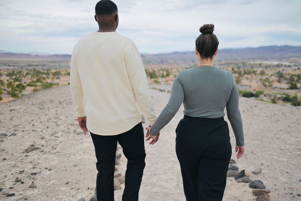Couple holding hands as they walk away on a rocky desert trail overlooking a sparse valley and distant mountains under a cloudy sky.