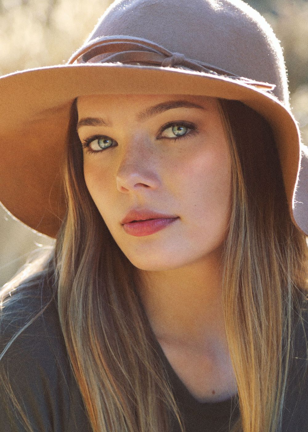 Close-up portrait of a young woman with green eyes and long hair wearing a tan wide-brimmed felt hat in warm sunlight.