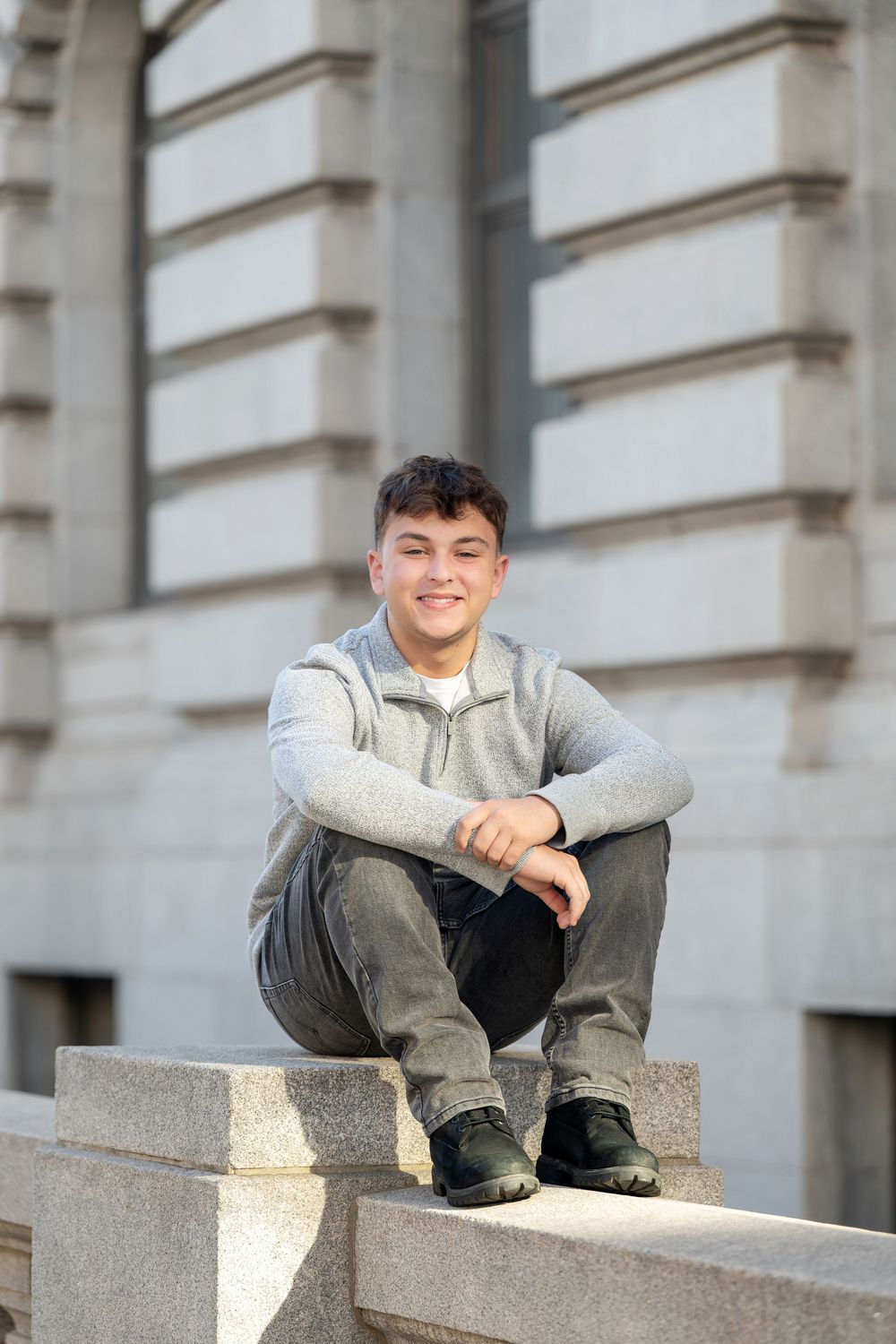 Smiling teenage boy in a gray quarter-zip sweater and black boots sits on a stone ledge in front of a classic stone building.