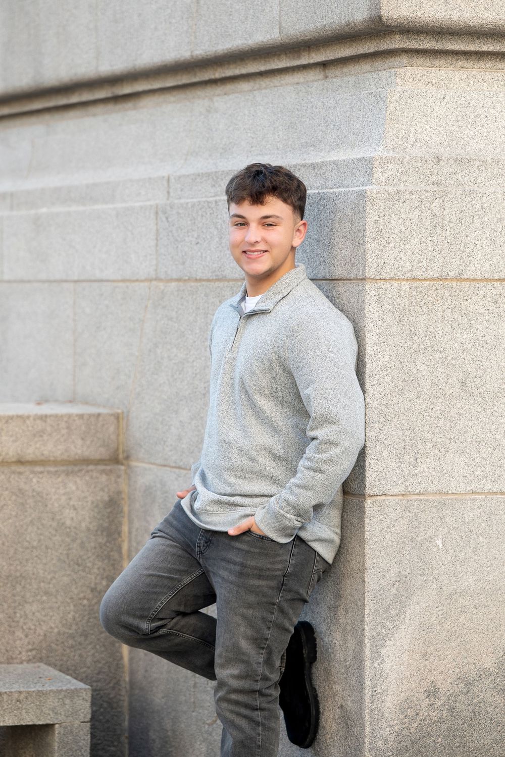 Smiling young man in a gray quarter-zip sweater and jeans leans casually against a large stone building wall with one foot propped up.