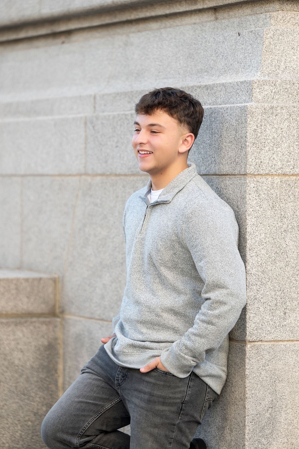 Smiling teenage boy in a gray quarter-zip sweater leans against a granite stone wall with his hands in his pockets.