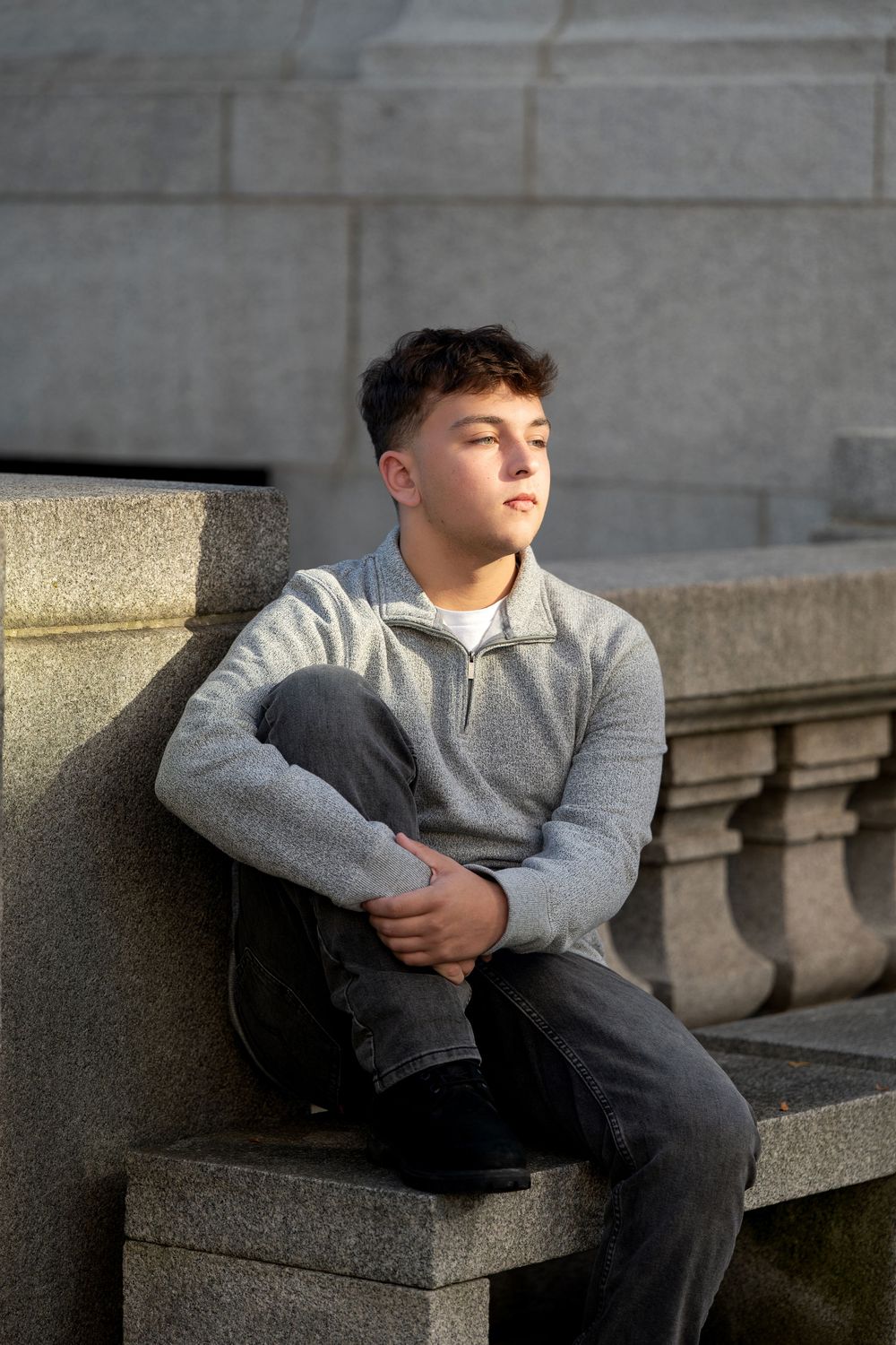 Teenage boy in a gray quarter-zip sweater and dark jeans sits on a granite ledge, gazing into the distance in warm sunlight.