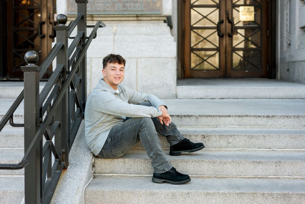 Smiling teenage boy in a gray sweatshirt and jeans sits on stone steps outside a building with ornate wooden doors and iron railings.