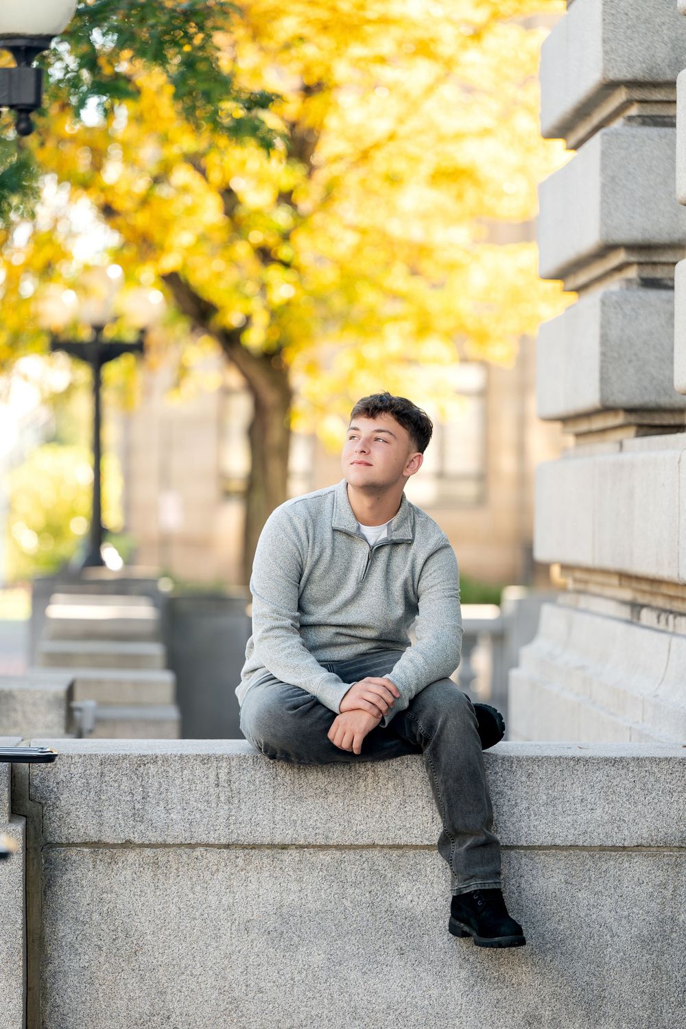 Young man in a gray quarter-zip sweater sits on a granite ledge, gazing upward near a golden autumn tree and a vintage lamppost.
