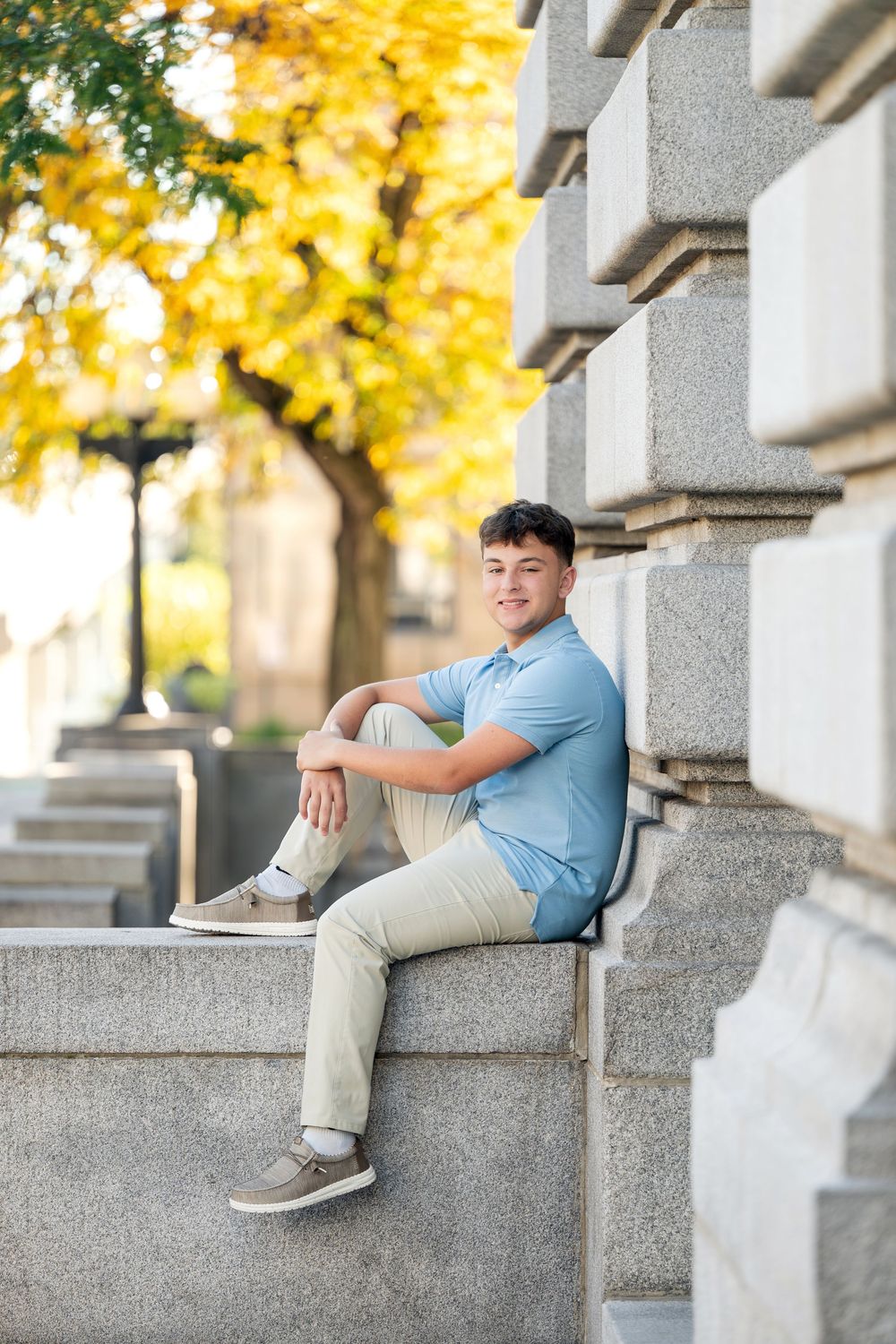 Young man in a light blue polo and khaki pants sits smiling on a stone ledge with golden autumn foliage behind him.