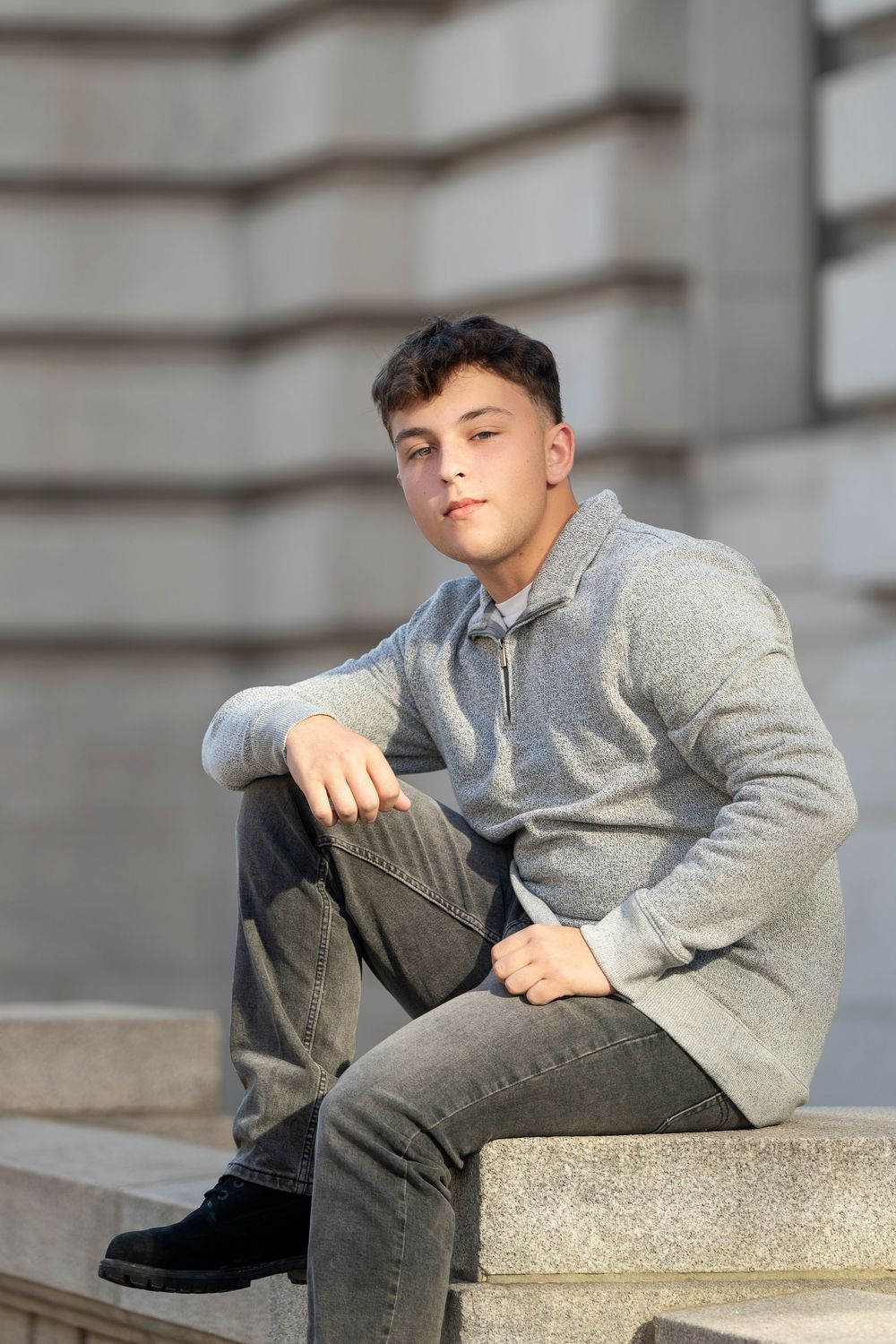Teenage boy in a gray quarter-zip sweater and jeans sits on stone steps in front of a classical building during a senior portrait session.