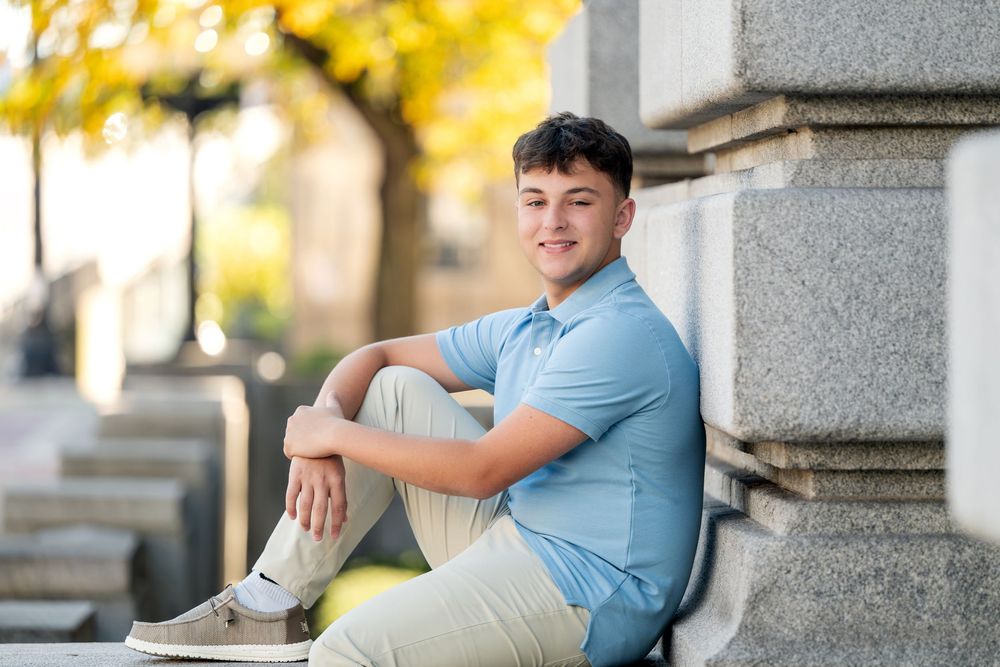 Young man in a light blue polo and khaki pants sits smiling on granite steps beside stone pillars with golden autumn trees behind him.