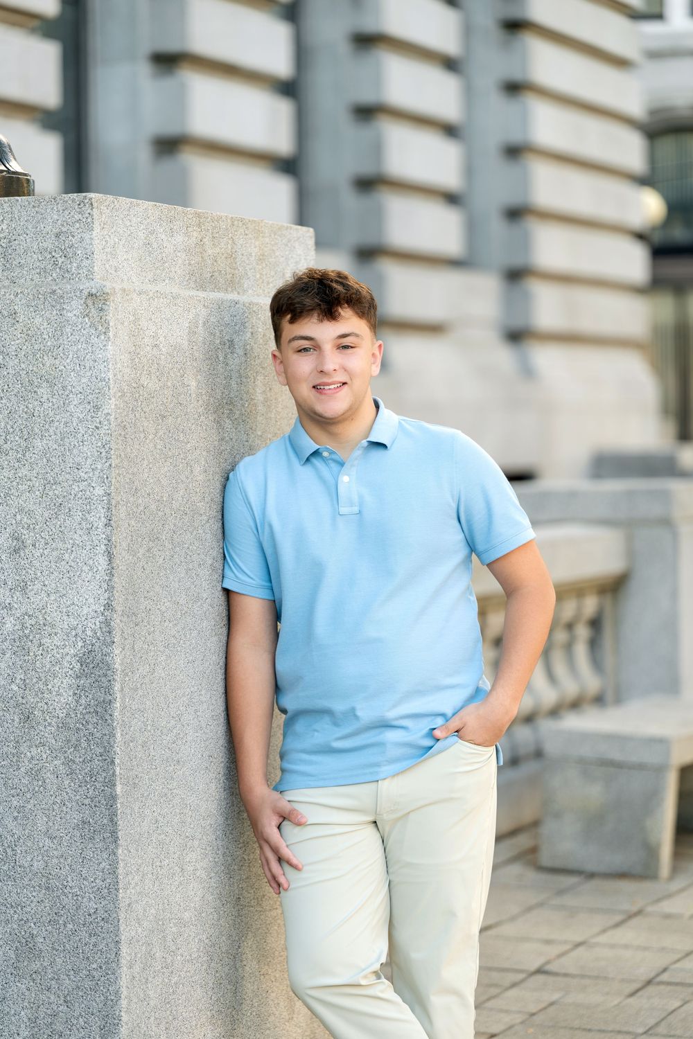 Smiling teenage boy in a light blue polo and khaki pants leans casually against a granite pillar outdoors near a stone building.