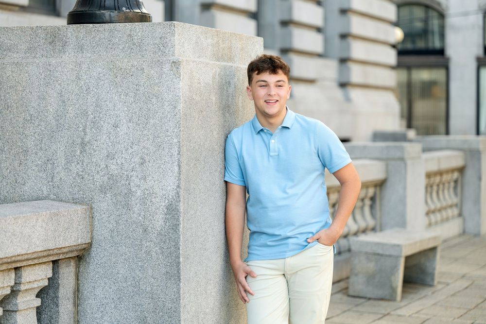 Smiling young man in a light blue polo and khaki pants leans against a stone pillar beside a balustrade outside a classic building.