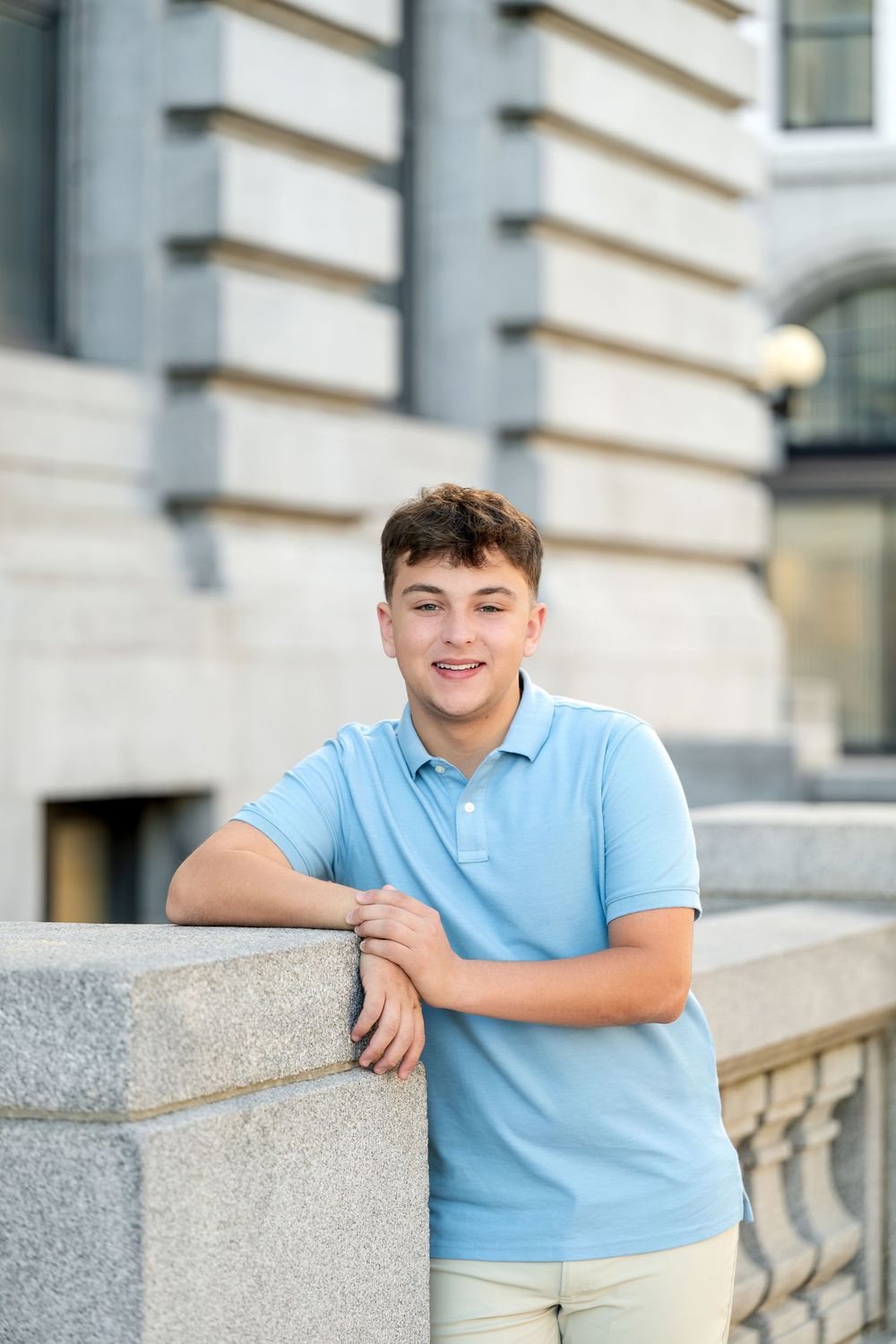 Smiling teen in a light blue polo leans on a granite wall with a balustrade and historic stone architecture behind him.