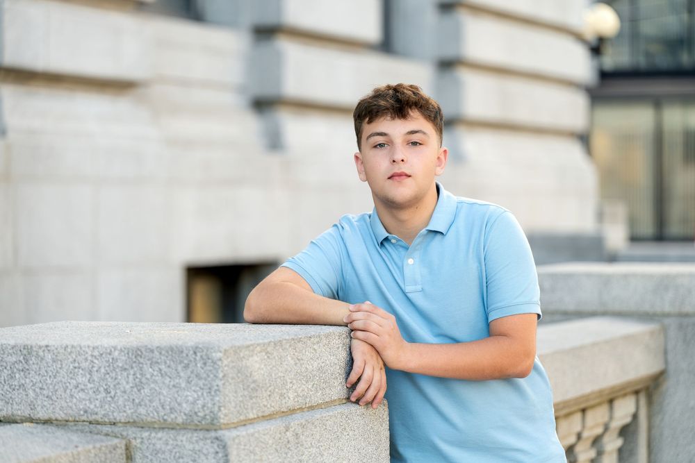 Teenage boy in a light blue polo leans on a stone ledge with arms crossed, posing outdoors beside a classic stone building.