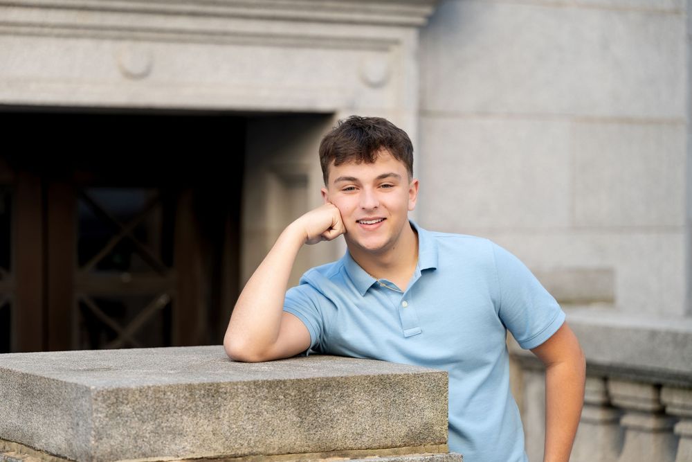 Smiling young man in a blue polo rests his chin on his fist while leaning on a stone ledge in front of a historic building.