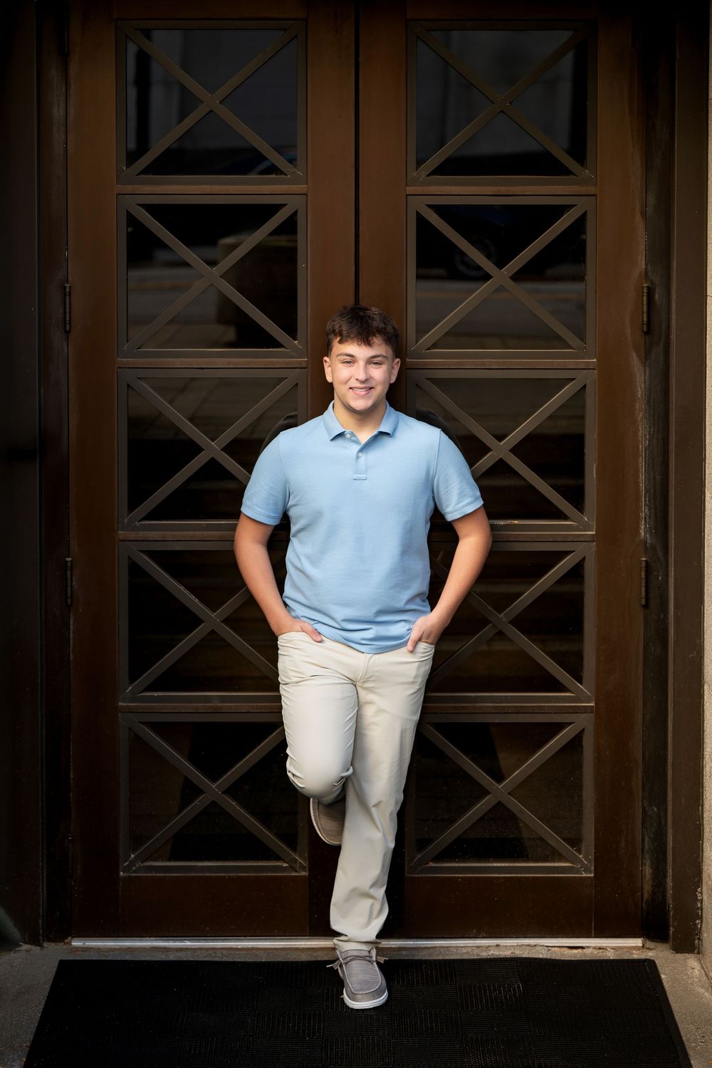 Smiling young man in a light blue polo and khaki pants leans in a dark wooden doorway with geometric glass panels, hands in pockets.