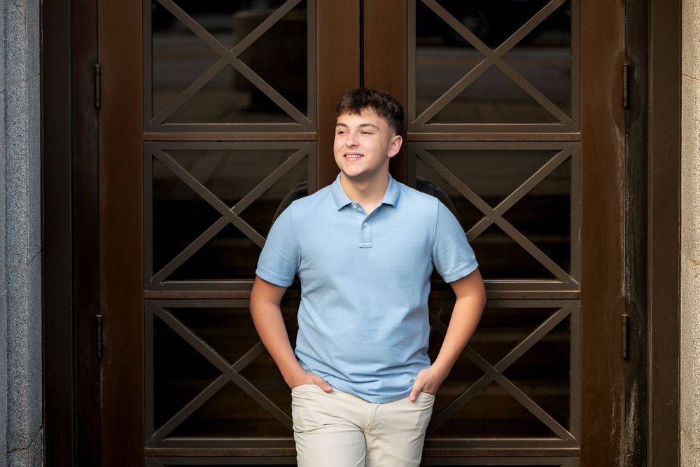 Young man in a light blue polo with hands in pockets stands before tall wooden doors with decorative diamond-patterned glass panes.