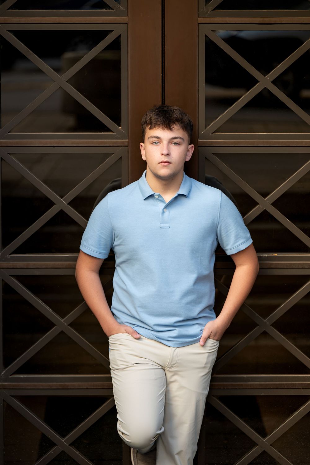 Young man in a light blue polo and khaki pants leans against ornate dark wood and glass doors with his hands in his pockets.