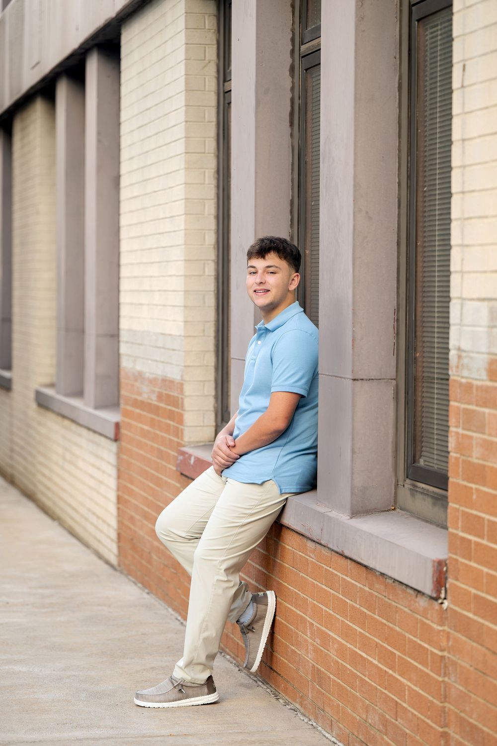 Smiling young man in a blue polo, khakis and gray sneakers leans on a window ledge of a red brick building in a senior portrait.