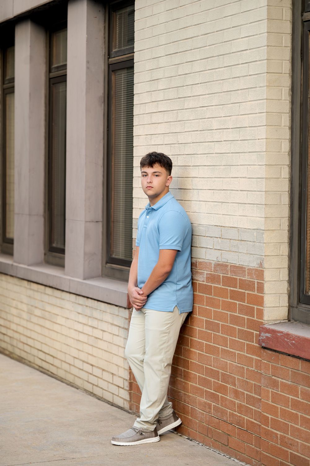 Young man in a light blue polo and khaki pants leans against a cream and red brick wall, gazing off to the side on a city sidewalk.