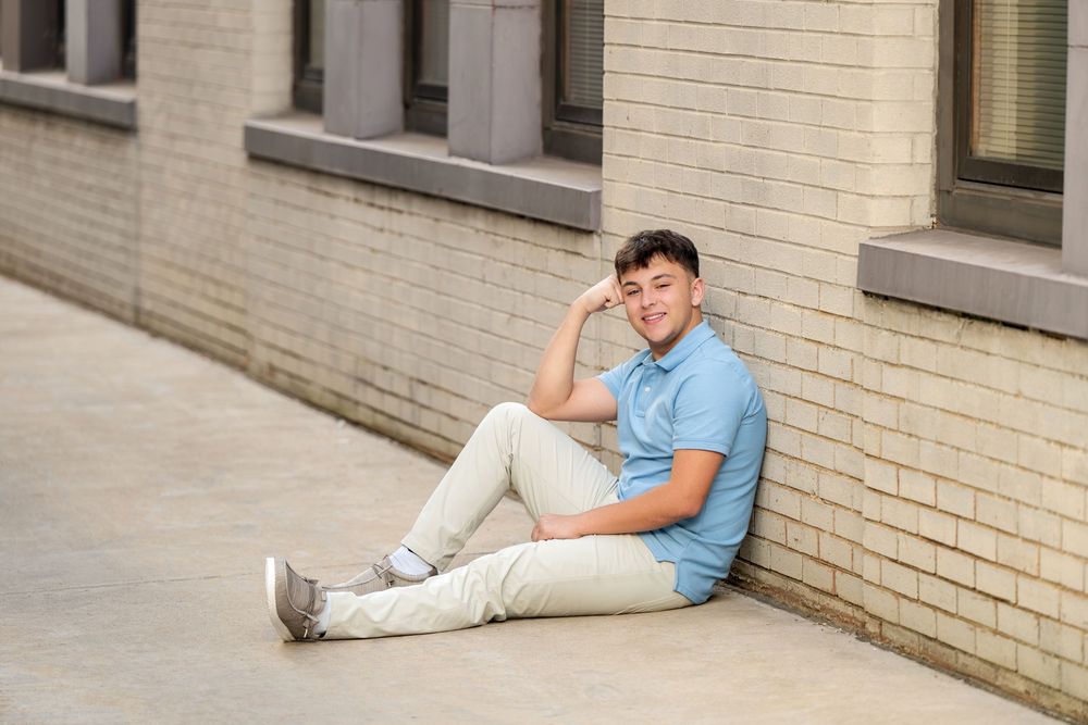 Young man in a blue polo and khaki pants rests his head on his hand while sitting against a light brick building wall.