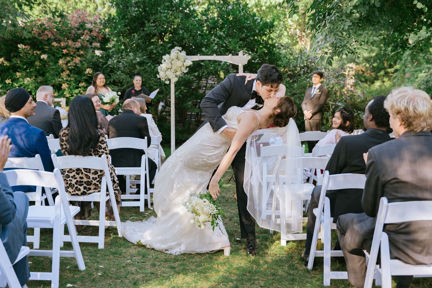 Bride and groom share a romantic dip kiss at outdoor garden wedding ceremony with white chairs and floral arch.