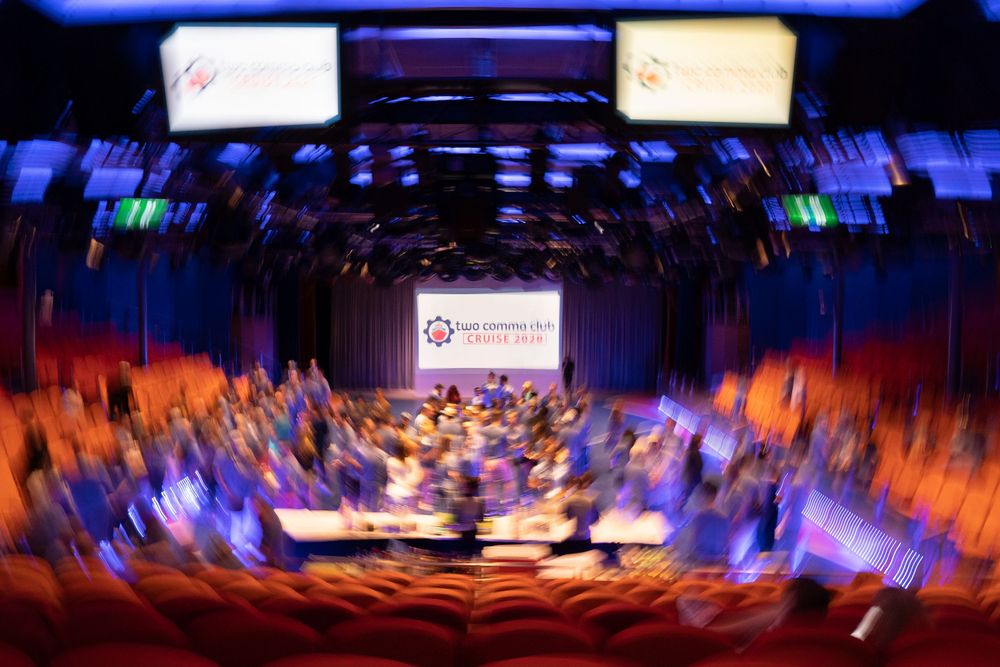 Blurred motion shot of a packed conference hall with blue lighting, large screens, and rows of attendees at a corporate event.