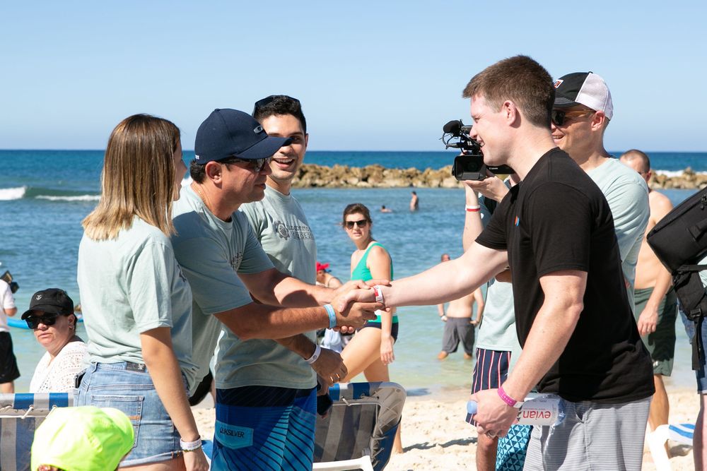 People shaking hands on a sunny beach while a cameraman films the outdoor event near the ocean.