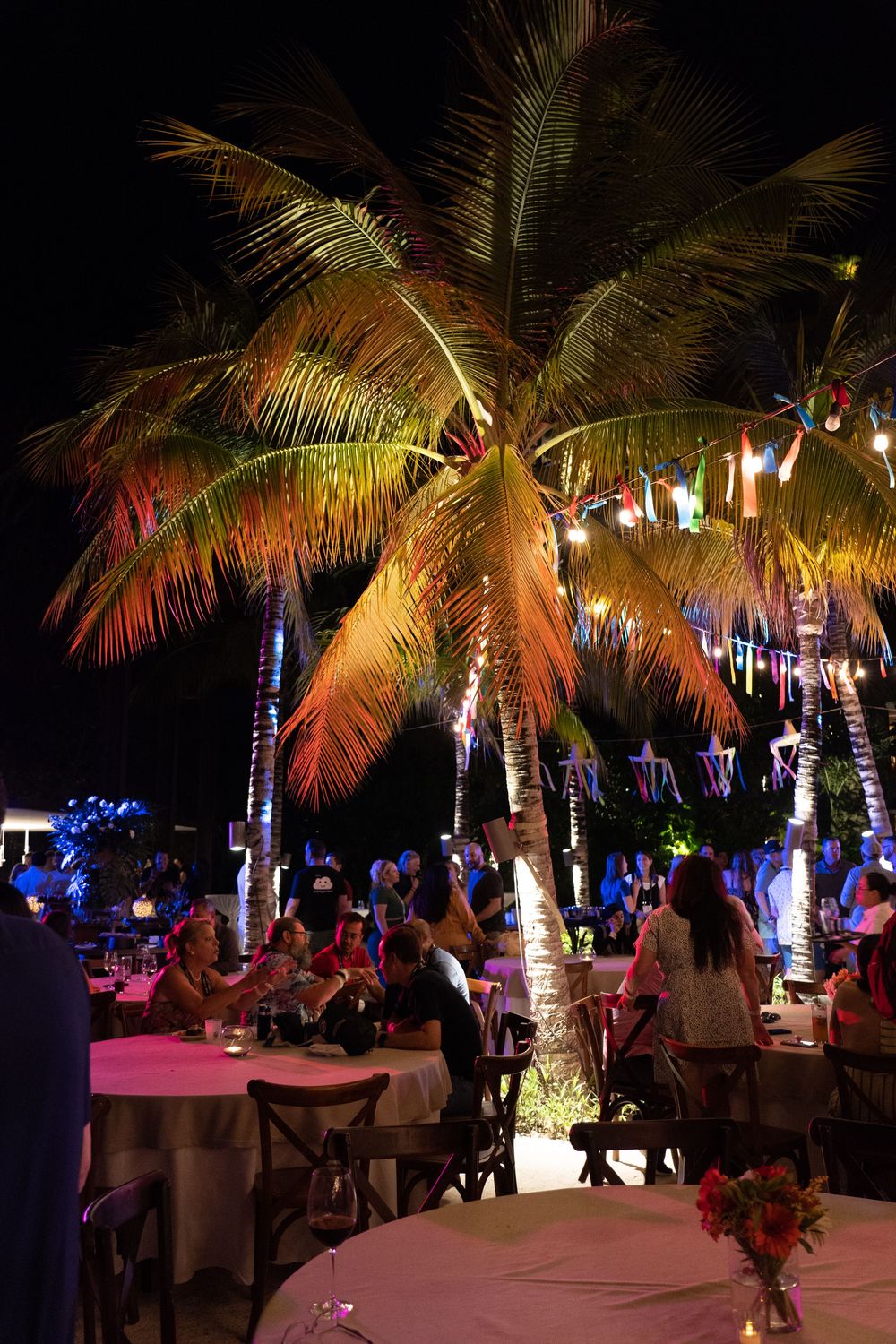 Tropical outdoor dining event at night with illuminated palm trees, colorful lights, and guests seated at white-clothed tables.