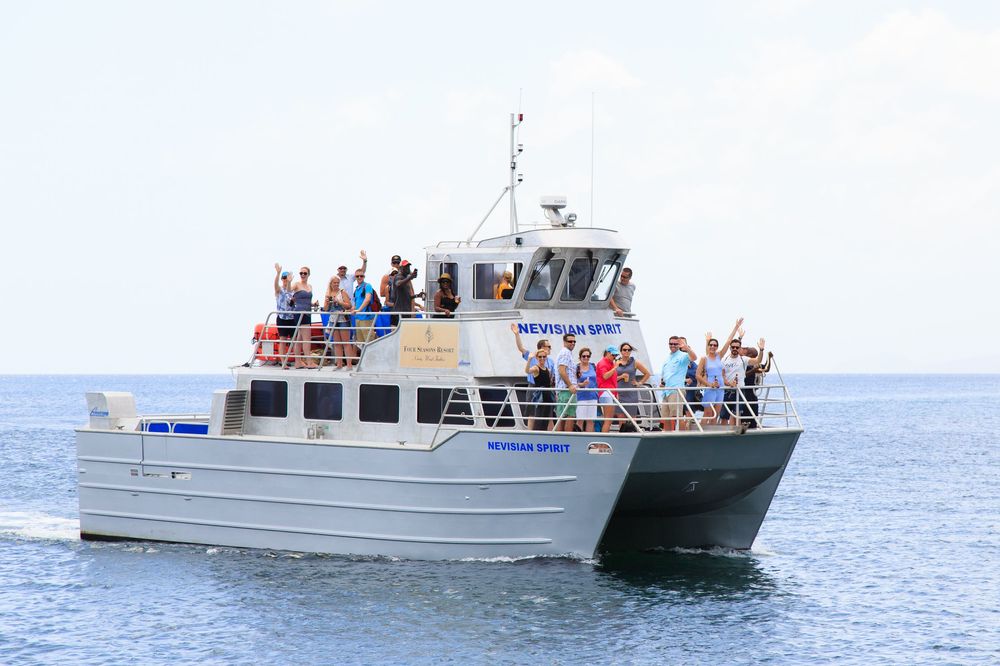 A crowded charter boat filled with waving tourists cruises on calm blue ocean waters under a partly cloudy sky.
