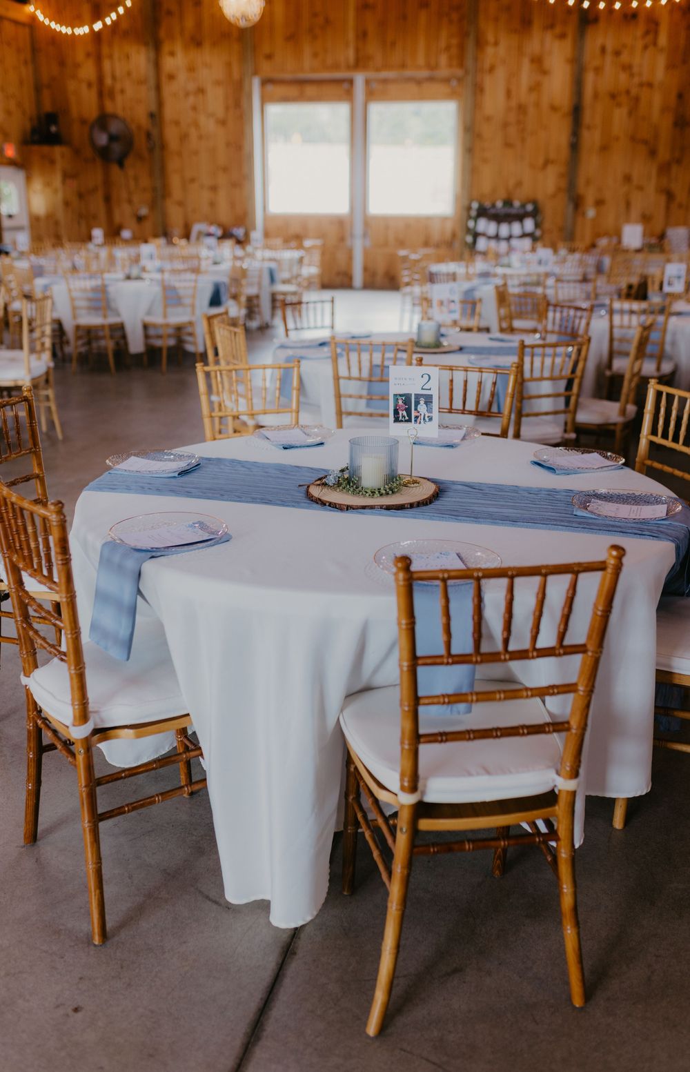 Rustic barn wedding reception with round tables, white linens, blue runners, gold Chiavari chairs, and elegant centerpieces.