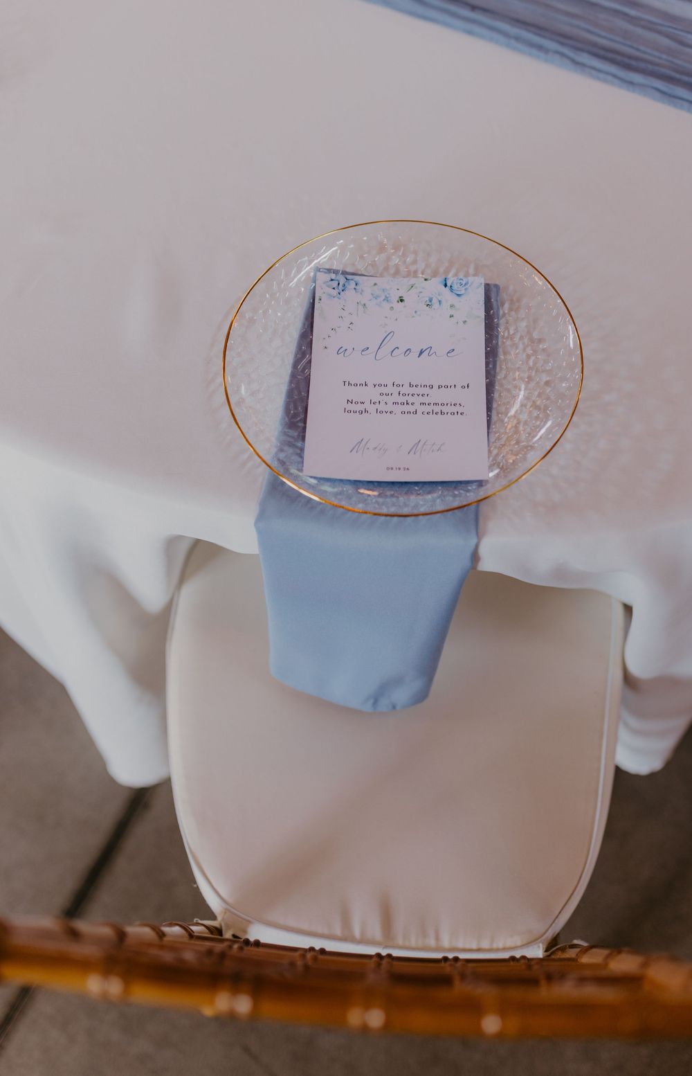 Elegant wedding place setting with blue napkin, gold-rimmed plate, and welcome card on a chiavari chair.
