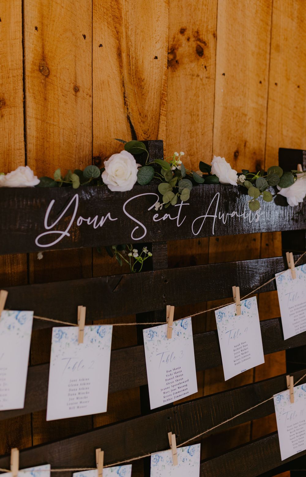 Wedding seating chart display with clothespin cards on twine, white roses, greenery, and "Your Seat Awaits" sign on dark wood.