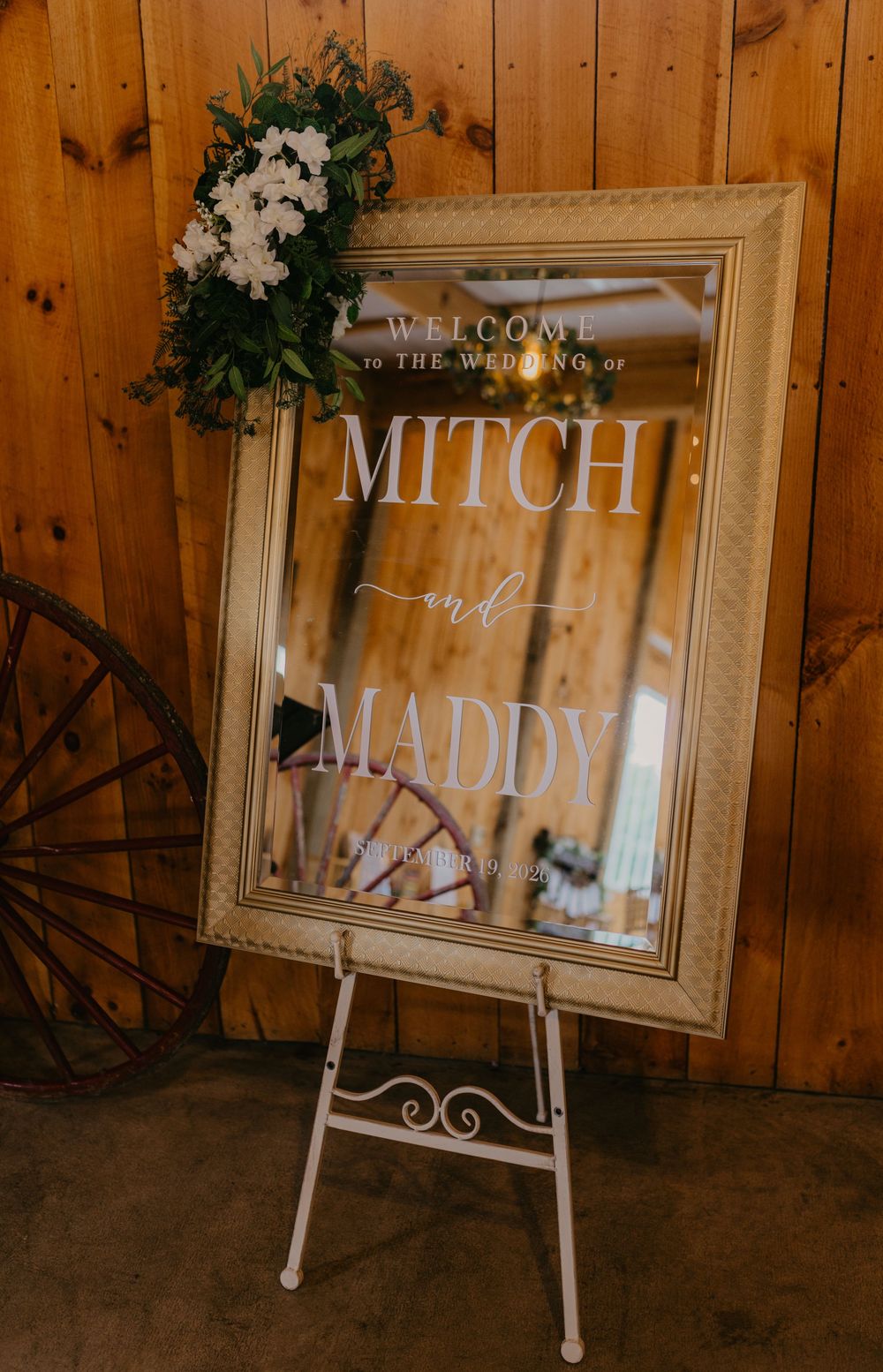 Elegant wedding welcome mirror sign for Mitch and Maddy on white easel with floral decor in rustic barn venue.