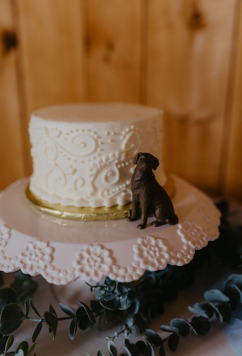White wedding cake with lace piping and chocolate Labrador dog topper on pink stand with eucalyptus.