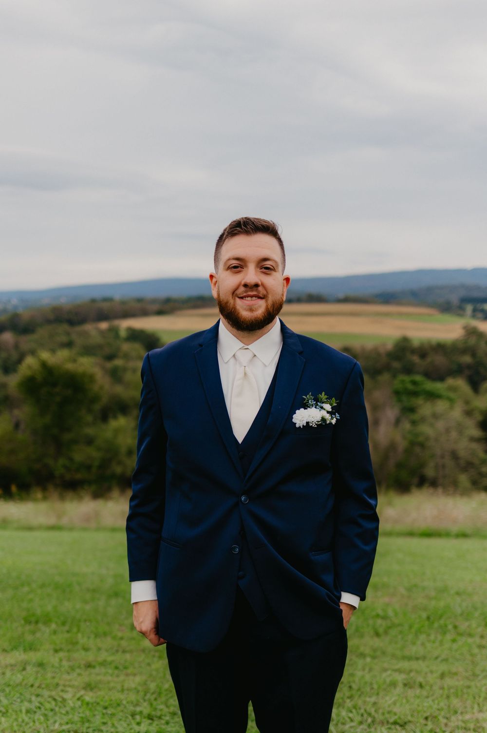 Smiling groom in navy blue suit with white boutonniere stands on green lawn with rolling hills backdrop.