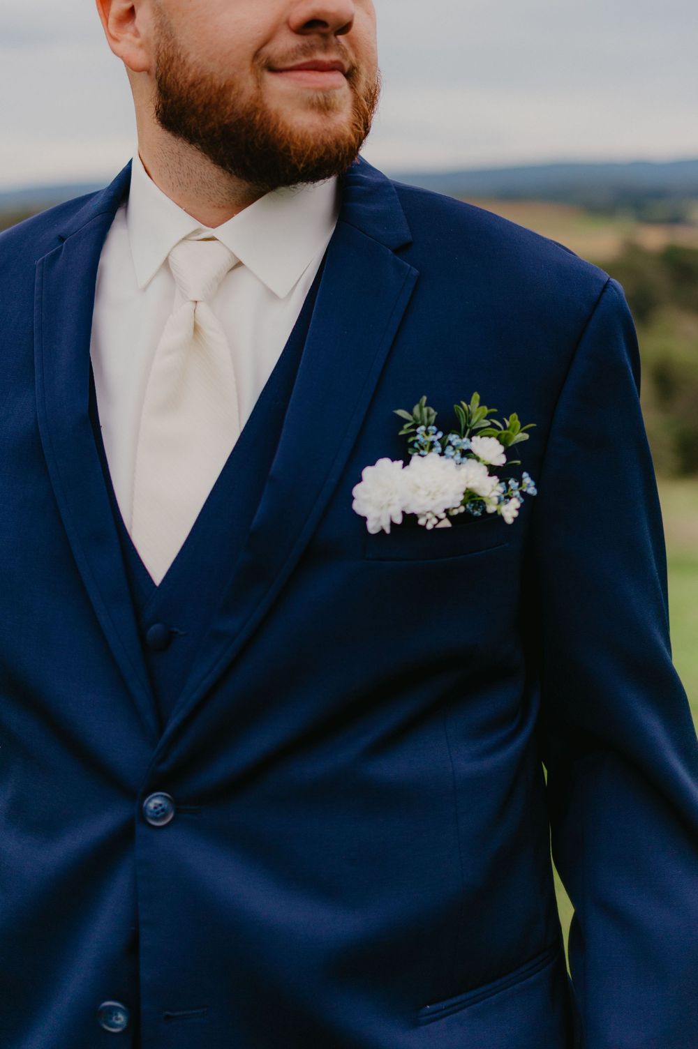 Groom in navy blue three-piece suit with cream tie and white floral pocket square boutonniere at outdoor wedding.