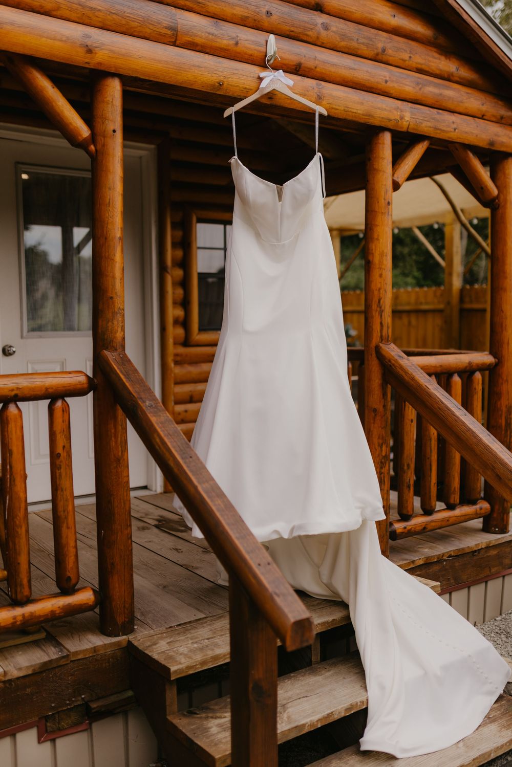 Elegant white satin wedding dress with train hanging on a hanger outside a rustic log cabin porch.