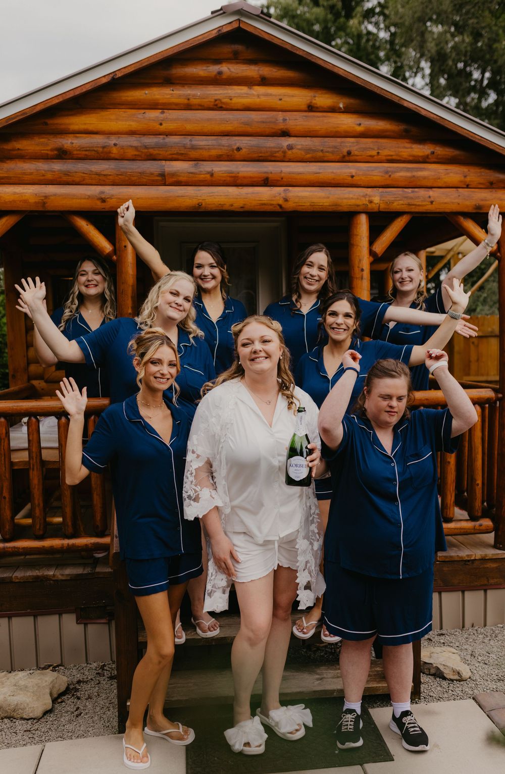 Bride in white robe holds champagne with bridesmaids in navy pajamas celebrating outside a rustic log cabin.
