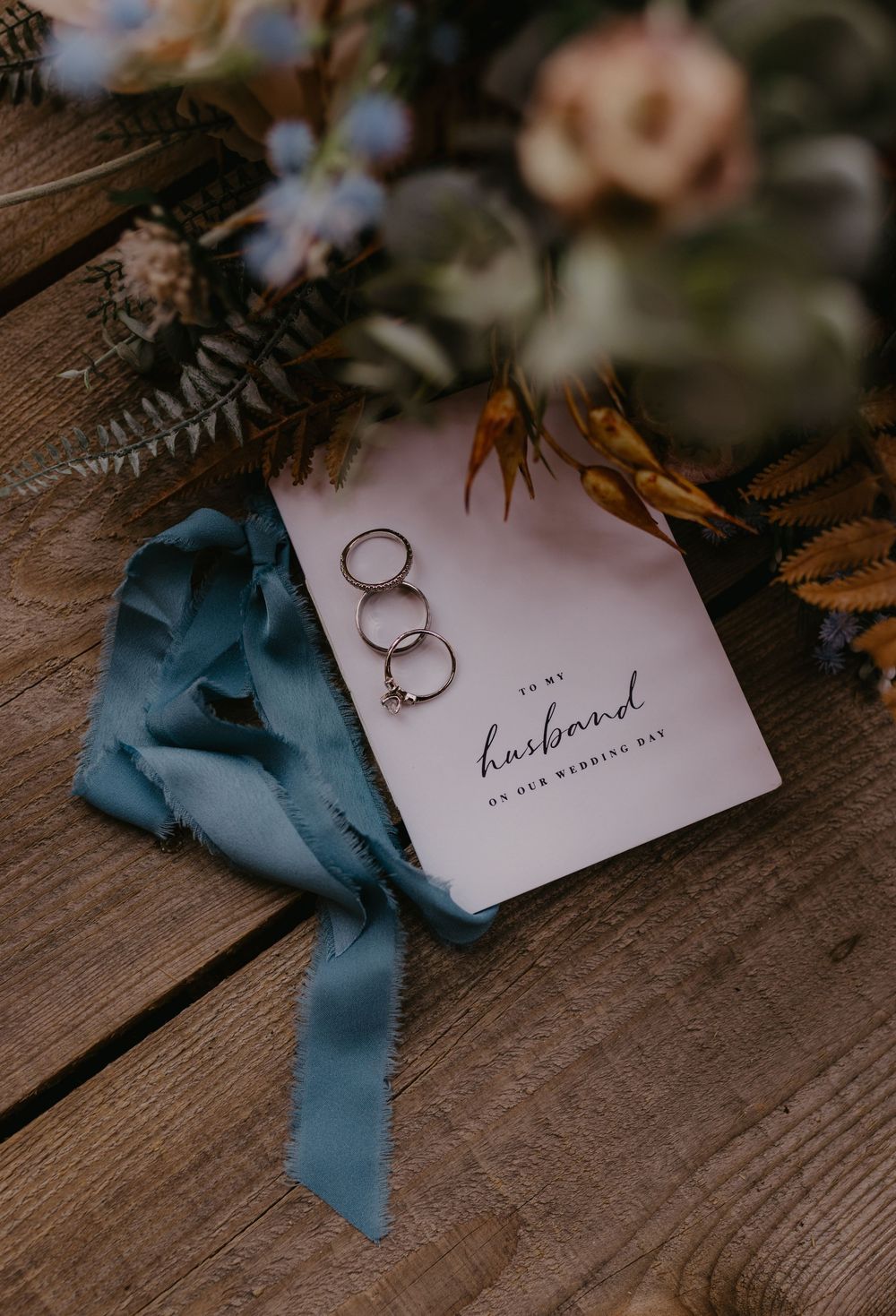 Wedding rings resting on a husband card with blue ribbon and floral bouquet on rustic wood.