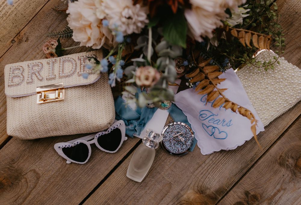 Bridal flat lay featuring a woven BRIDE purse, heart sunglasses, something blue charm, handkerchief, and floral bouquet on wood.