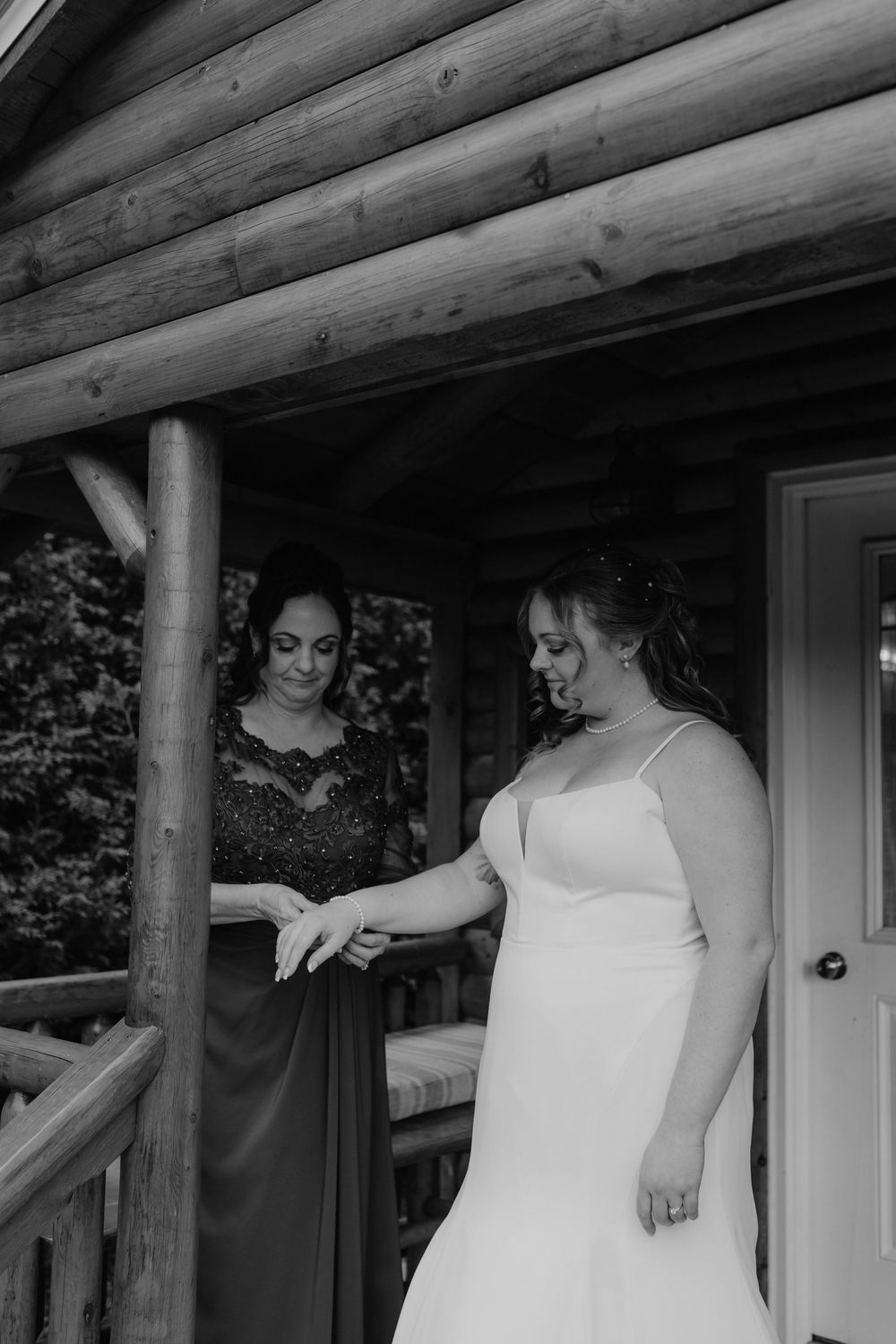 Bride in white gown getting ready on log cabin porch with bridesmaid in black lace dress, black and white wedding photo.