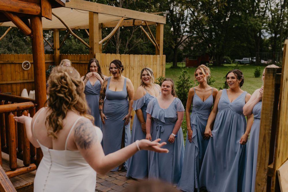 Bride in white gown reveals dress to bridesmaids in blue dresses outdoors at rustic wedding venue.